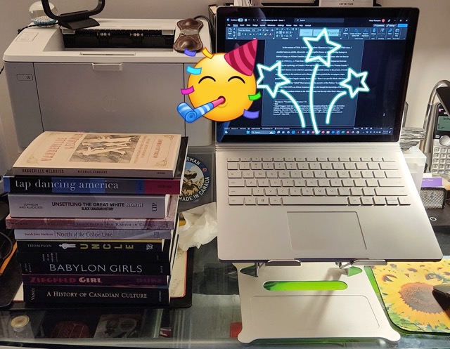 an open laptop with a celebratory smiley face emoji blocking the screen, a stack of books to the left of the computer 